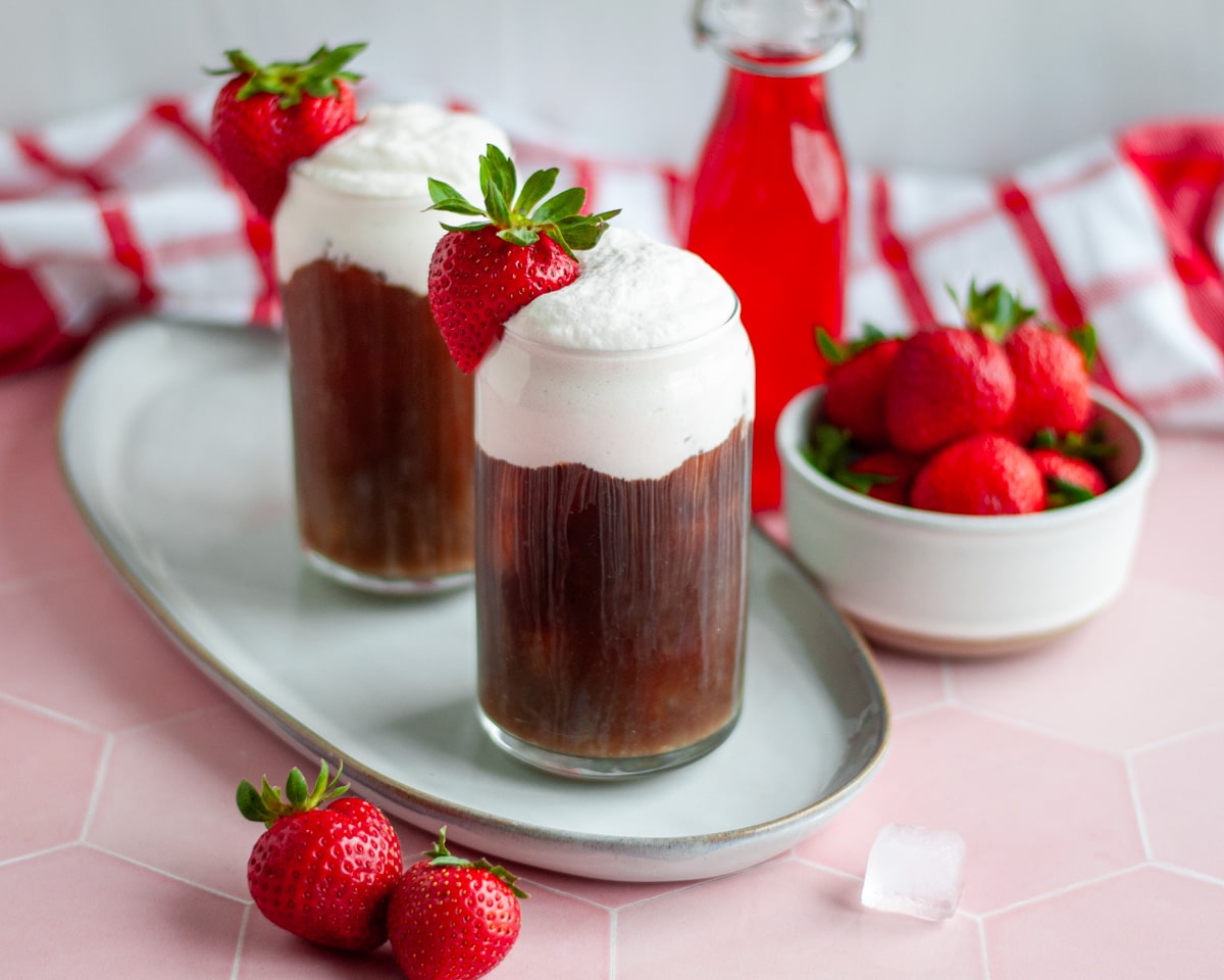 Two tall glasses on a tray filled with cold brew coffee topped with a generous portion of strawberry cold foam, a strawberry for garnish with more strawberries, strawberry syrup, and a checkered napkin around them.