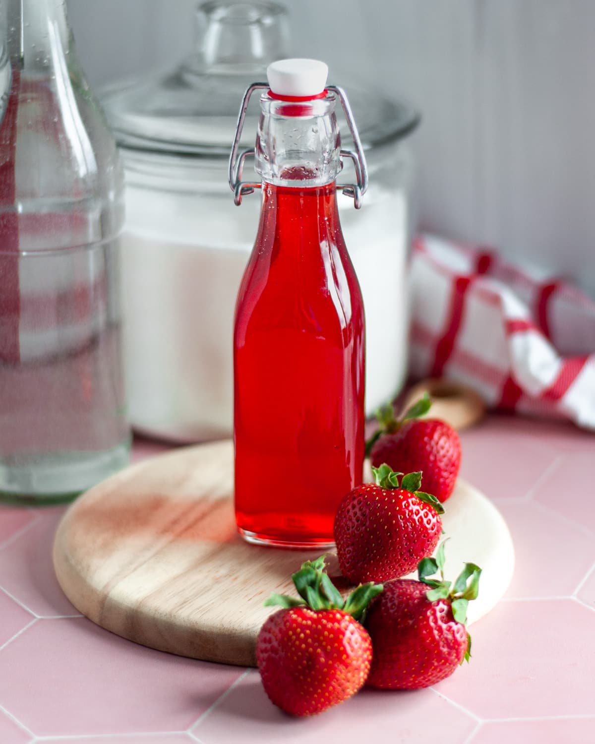 A lidded glass jar filled with strawberry simple syrup, sitting on a wooden board and surrounded by ingredients and a checkered linen.