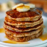 Side view of a stack of 5 freshly cooked banana pancakes on a white serving plate, topped with a few slices of bananas and maple syrup.