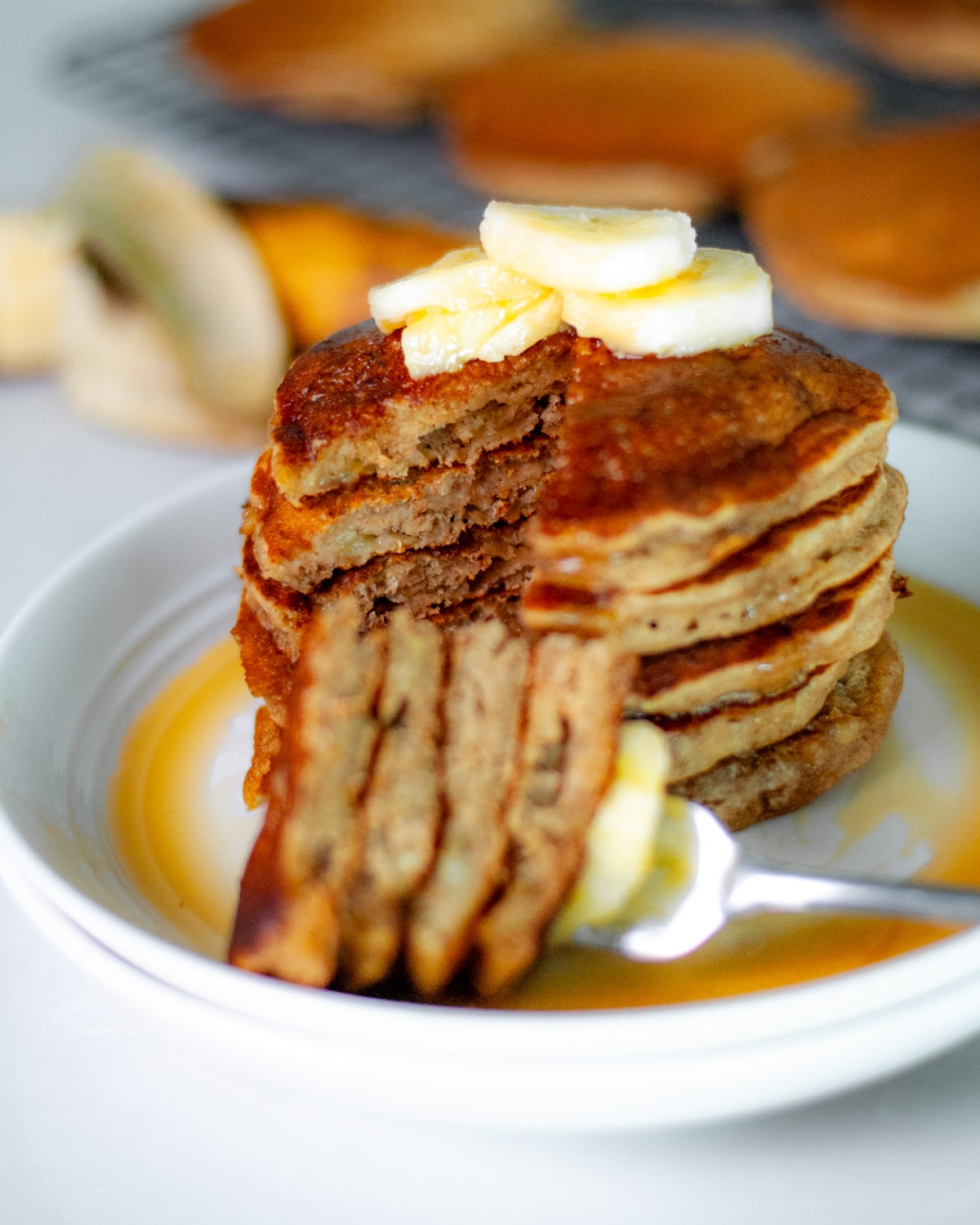 Close up of the inside of a stack of pancakes, with a fork-full of pancake slices set in front of the stack on a white serving plate, topped with a few slices of bananas and maple syrup.