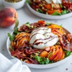 Process shot showing the arugula, peach slices, burrata, crispy prosciutto, and pistachio pieces arrange on a serving plate with an olive oil and balsamic glaze drizzle on top.