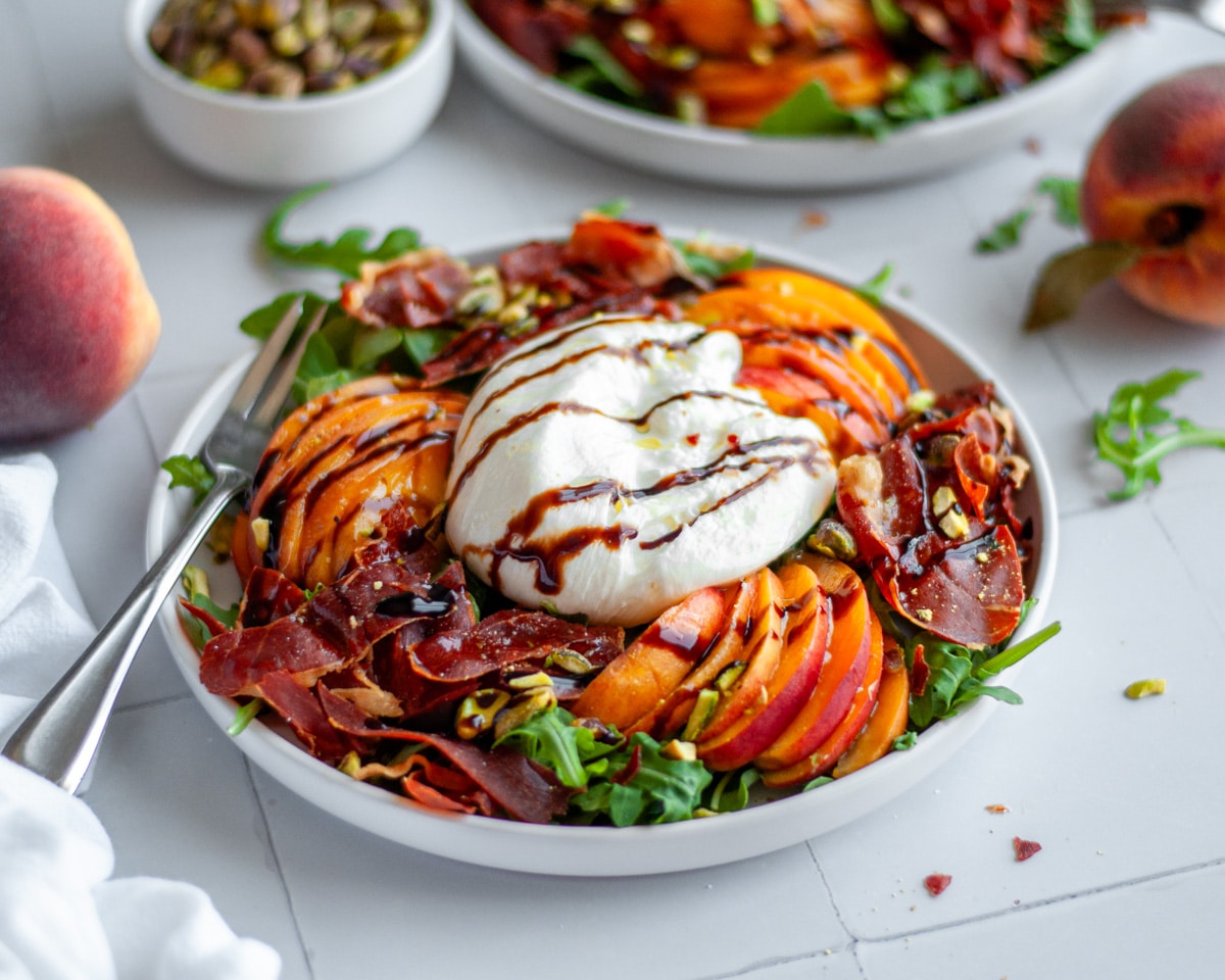 Two plates of peach arugula salad with burrata and crispy prosciutto, topped with an olive oil and balsamic glaze drizzle, with additional ingredients scattered around them.