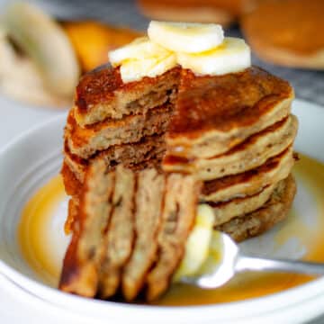 Feature image for our banana pancakes recipe, showing a close up of the inside of a stack of pancakes, with a fork-full of pancake slices set in front of the stack on a white serving plate, topped with a few slices of bananas and maple syrup.