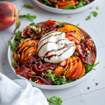 Two plates of peach arugula salad with burrata and crispy prosciutto, topped with an olive oil and balsamic glaze drizzle, with additional ingredients scattered around them.