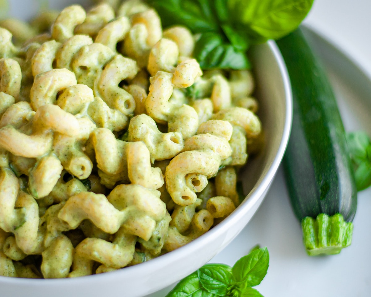 A white serving bowl of zucchini pasta, with a fresh zucchini and additional basil next to it.