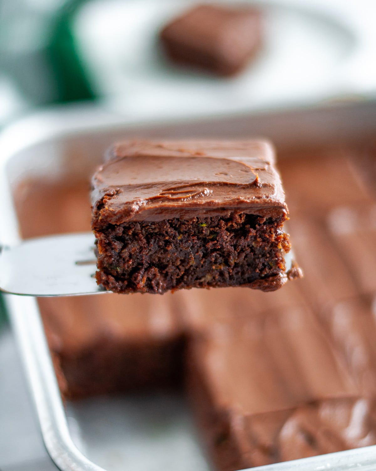 Close up of a chocolate zucchini brownie with frosting on a metal spatula in front of the pan of brownies.