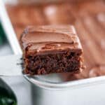 Close up of a chocolate zucchini brownie with frosting on a metal spatula in front of the pan of brownies.
