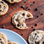 Close up of an eggless chocolate chip cookie with a bite out of it, surrounded by chocolate chips, a wire cooling rack filled with cookies, and a plate of cookies.