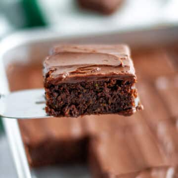 Feature image showing a close up of a chocolate zucchini brownie with frosting on a metal spatula in front of the pan of brownies.