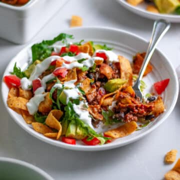 Close up of a white serving plate with a scoop of walking taco casserole topped with lettuce, tomatoes, avocado, and sour cream, with a fork holding a bite.