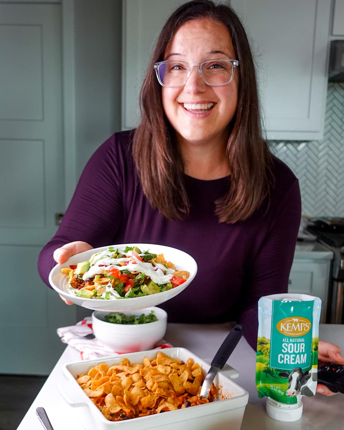 Meredith holding the a plate filled with a scoop of walking taco casserole towards the camera, topped with lettuce, tomatoes, avocado, and sour cream.