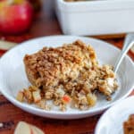 Close up of a slice of baked apple oatmeal on a white plate with a fork with the pan of baked oatmeal, an apple, oats, apple slices, and a jar of applesauce behind it.