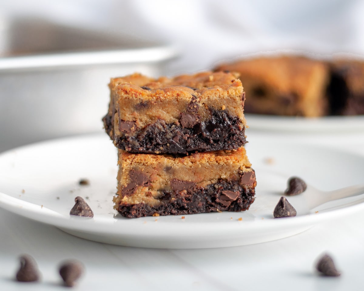 Two brookie bars stacked on top of each other on a white serving plate, with chocolate chips, crumbs, and more brookies around it.