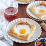 Close up of puff pastry egg tarts on white scalloped serving plates, with a white linen, a bowl of fruit salad, and a glass of rose wine behind it.