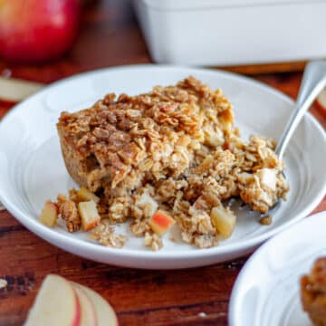 Feature image showing a close up of a slice of baked apple oatmeal on a white plate with a fork with the pan of baked oatmeal, an apple, oats, apple slices, and a jar of applesauce behind it.