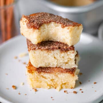 Feature image of a stack of three snickerdoodle bars, with the top bar having a bite out of it, on a serving plate with the pan of bars and cinnamon sticks in the background.