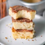 A stack of three snickerdoodle bars, with the top bar having a bite out of it, on a serving plate with the pan of bars and cinnamon sticks in the background.